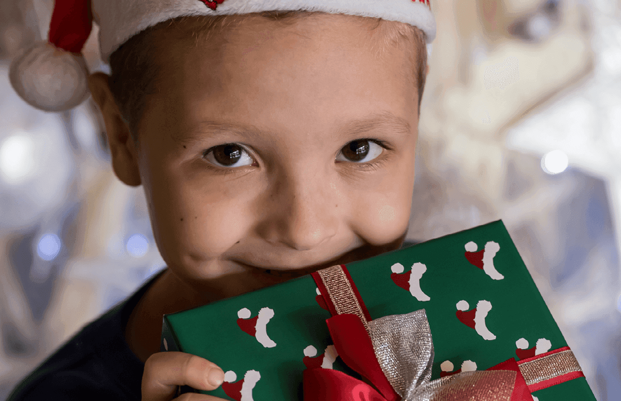 School-age boy smiling at the camera while wearing a Santa hat and hiding half of his face behind a gift