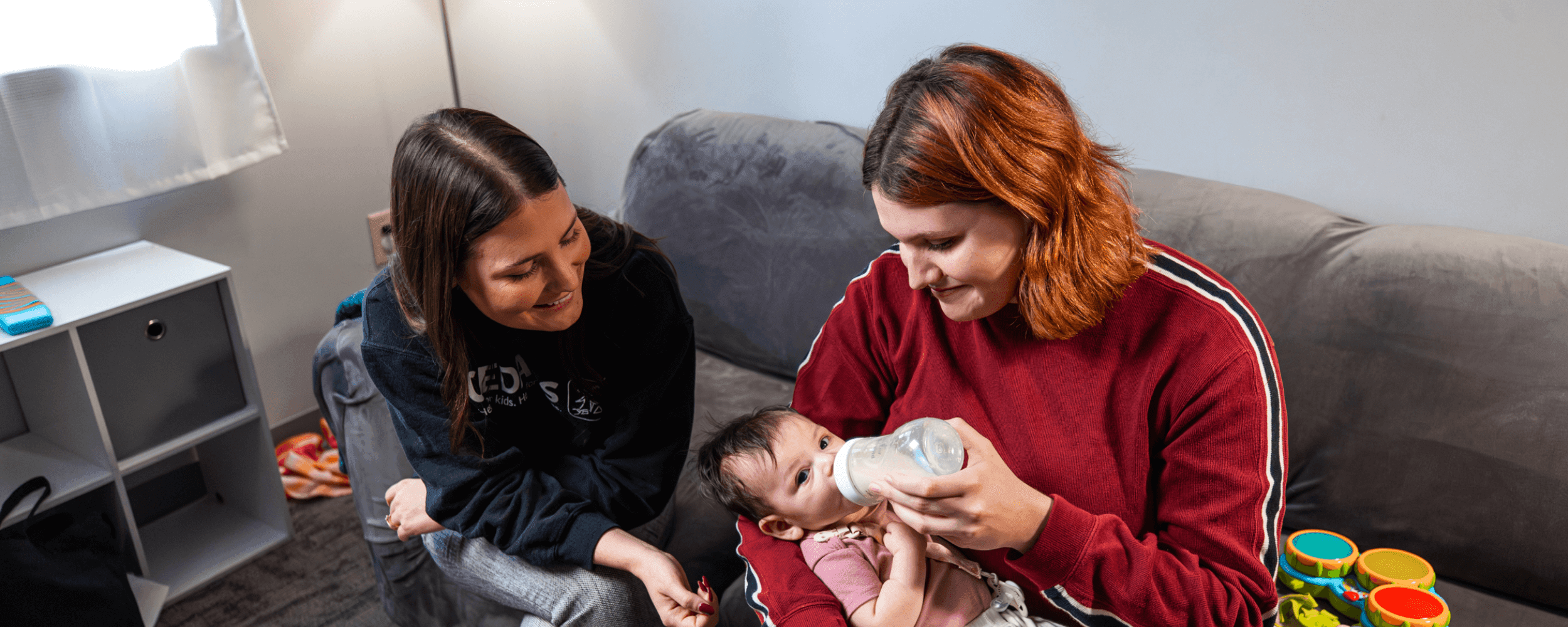 Young mom feeding her baby a bottle while a CEDARS staff member helps