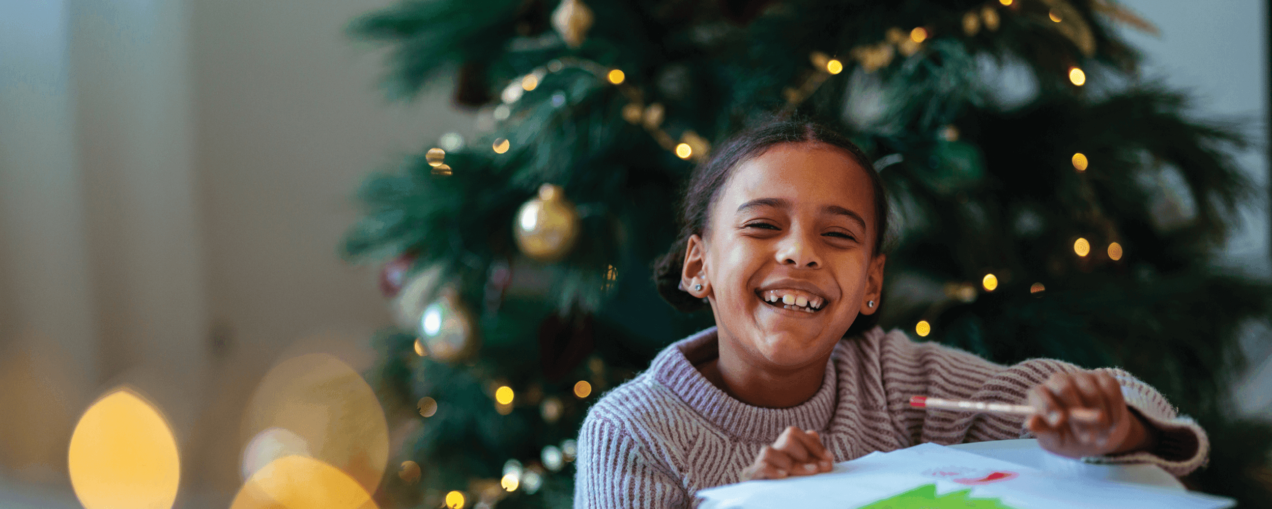 School aged girl smiling big while drawing next to a Christmas tree.