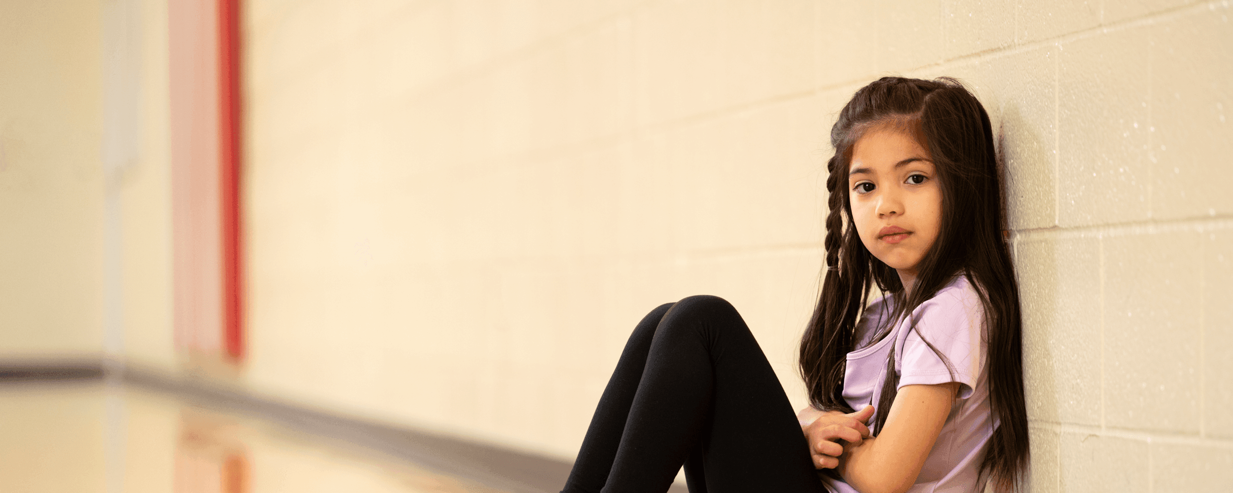 School age girl sitting on the floor of a school gym and somberly looking at the camera