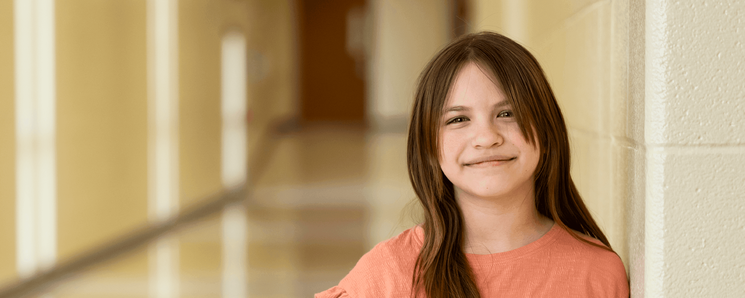 tween girl softly smiling and leaning against the wall in a school hallway 